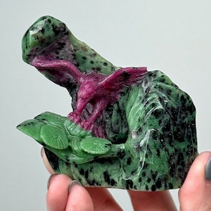 Puede incluir: Una escultura de piedra tallada que presenta un águila de color magenta intenso con las alas extendidas posada sobre una base moteada de verde y negro. El águila está situada sobre dos formas circulares planas de color verde.