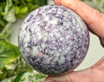 May include: A spherical gemstone with a textured surface, showcasing a blend of white and purple hues. The orb-shaped stone has a crystalline appearance, with small pink inclusions. The image is a close-up, highlighting the intricate details of the polished crystal.