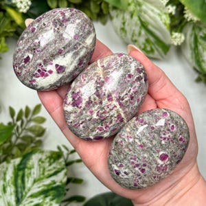 May include: Three oval-shaped stones with a mix of gray, white, and pink hues. The stones have a speckled appearance, with the pink color concentrated in small, irregular patches. The stones are smooth and polished, with a natural, organic look.