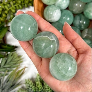 May include: Three green fluorite spheres, each with a different pattern of white veins, are held in a hand. The spheres are smooth and polished.