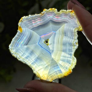 May include: A translucent, irregularly shaped agate slice. The stone features concentric bands of white, blue, and iridescent colors, with a yellow and orange outer edge. The agate is held by a person's fingers, showcasing its natural beauty.
