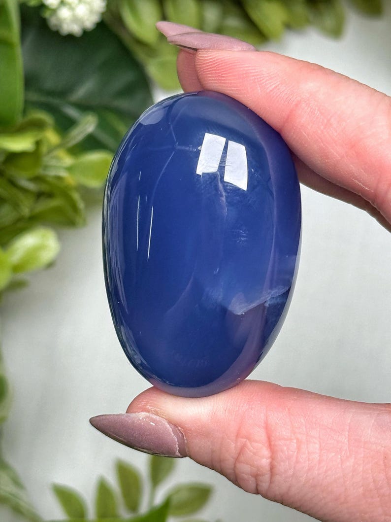 May include: A smooth, oval-shaped, deep blue gemstone is held between fingers. The polished stone has a glossy surface with subtle white veining. The background features green foliage. The gemstone is approximately 2 inches in length.