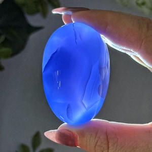 May include: A translucent, oval-shaped blue gemstone is held between two fingers. The stone has a smooth, polished surface with internal veining and a slight crack. The gemstone is illuminated, creating a glowing effect.