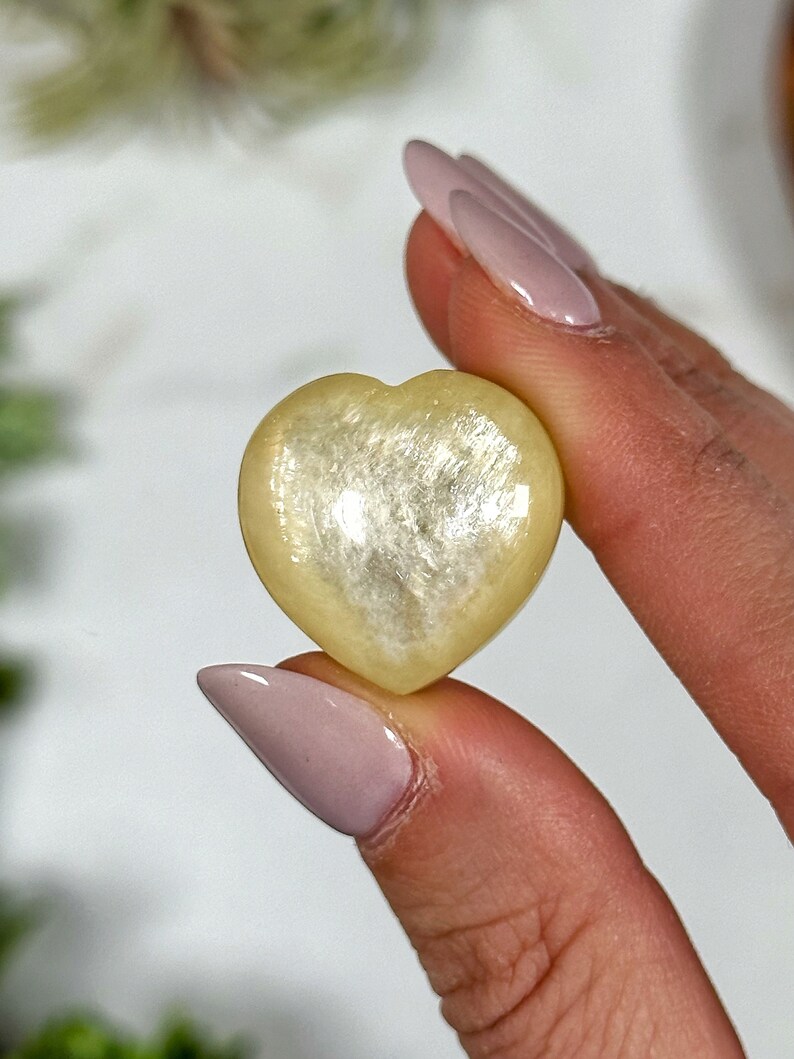 May include: A heart-shaped, pale yellow selenite crystal with a shimmering, iridescent finish.