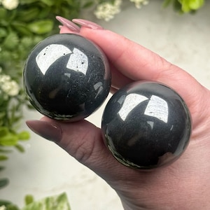 Könnte beinhalten: Zwei polierte, dunkelgraue Kugeln werden in einer Hand gehalten. Die Kugeln haben eine reflektierende, glänzende Oberfläche mit hellen weißen Reflexen. Der Hintergrund umfasst grünes Laub und weiße Blüten. Diese Kugeln sind wahrscheinlich dekorative oder Sammlerobjekte.