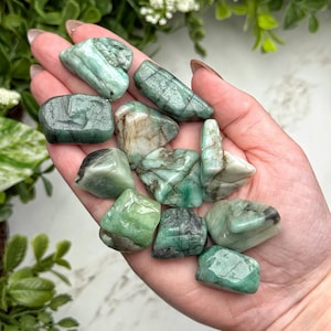May include: A handful of polished emerald gemstones in varying shapes and sizes. The stones display a range of green hues, from light to deep emerald, with some showing natural inclusions. The gemstones are held in an open hand against a backdrop of greenery.