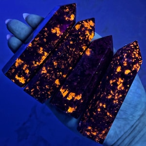 May include: Four dark purple crystals with orange speckles are held in a hand. The crystals are rectangular with pointed tops. The crystals are illuminated with a blue light, creating a glowing effect.