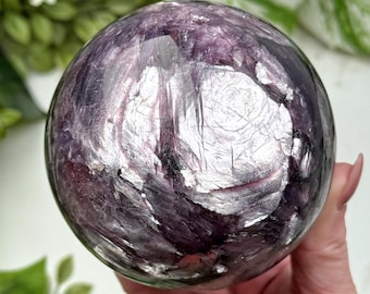 May include: A polished sphere of purple and silver stone. The orb has a reflective, layered appearance with a mix of dark purple and shimmering silver tones. The sphere is held in a hand, with green foliage in the background.