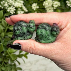 May include: Two small snail-shaped carvings made from green zoisite stone with black inclusions and small patches of deep red ruby. The carvings feature a smooth, polished surface and are shaped to resemble the coiled shell and head of a snail.