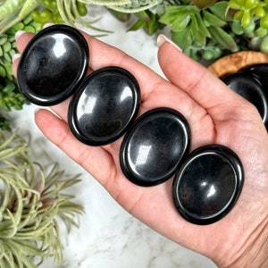 May include: Four smooth, black obsidian palm stones, also known as worry stones, are held in a hand. The stones are oval shaped and polished to a high shine.