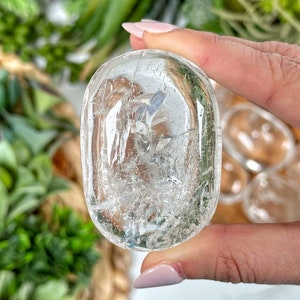 May include: A clear quartz crystal palm stone with a smooth, polished surface. The stone has a natural, cloudy appearance with visible inclusions.