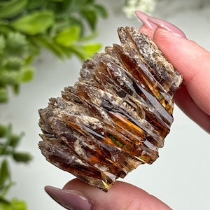 May include: A close-up of a rough, layered mineral specimen. The crystal formation features a mix of brown, amber, and clear sections, with a textured, crystalline appearance. The specimen is held in a person's hand, with a blurred green background.