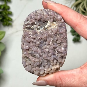 May include: A purple agate geode with a smooth, oval shape and a surface covered in small, round nodules. The geode has a natural, earthy look.