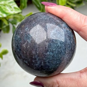 May include: A polished, dark blue sphere with a reflective surface. The sphere has a mottled appearance with shades of gray and hints of purple. The smooth surface reflects light, creating bright highlights. The sphere is held in a hand.
