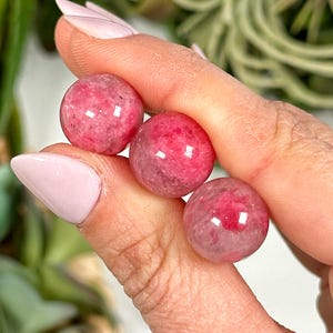 May include: Three pink and white gemstone spheres, each with a smooth, polished surface. The spheres are held in a hand, showcasing their translucent quality and natural variations in color.