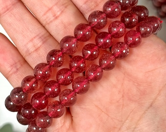 May include: Three strands of red gemstone bead bracelets. Each bracelet is composed of round, translucent beads with a deep red hue. The beads are strung on a red cord. The bracelets are displayed on a light background.