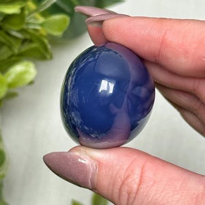 May include: A polished, oval-shaped gemstone in shades of blue and purple. The stone is held between fingers, showcasing its smooth, reflective surface. The gemstone's color gradient creates a captivating visual effect.