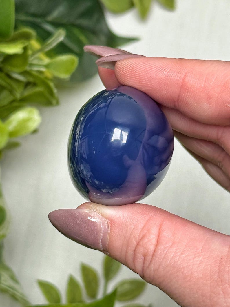 May include: A polished, oval-shaped gemstone in shades of blue and purple. The stone is held between fingers, showcasing its smooth, reflective surface. The gemstone's color gradient creates a captivating visual effect.