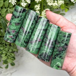 May include: Five cylindrical polished stones featuring a vibrant green base with speckled black inclusions and distinct patches of deep magenta-red. Each stone has a smooth, reflective surface and a uniform tubular shape, displaying natural mineral patterns.