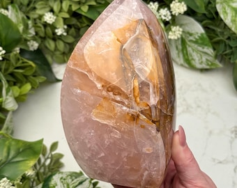 Op de afbeelding: Een gepolijst, druppelvormig kristal in roze en amber tinten. De steen heeft een glad, reflecterend oppervlak met natuurlijke adering en insluitsels. Het kristal wordt vastgehouden door een hand, met groen op de achtergrond.