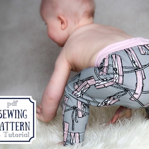 Può includere: Un bambino che indossa leggings grigi e rosa con un motivo a mollette. I leggings hanno una fascia rosa. Il bambino è in piedi su una superficie bianca e soffice. L'immagine include il testo "pdf SEWING PATTERN & Tutorial".