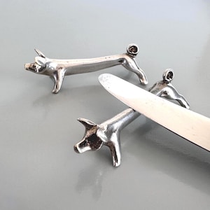 May include: Three silver-colored pig-shaped knife rests. The knife rests are in a lying down position, with the knife resting on top of one of the pig rests. The knife rests are on a gray surface.