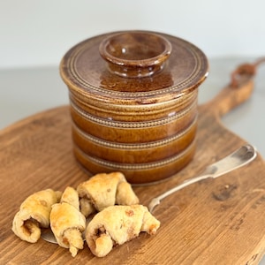 May include: A brown ceramic butter dish with a lid, featuring a decorative band design. The dish sits on a wooden cutting board with a silver butter knife and several crescent-shaped pastries.