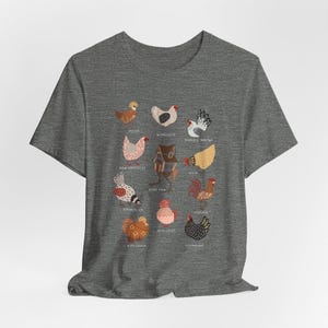 May include: A heather gray t-shirt featuring colorful illustrations of various chicken breeds. The shirt displays detailed drawings of chickens with names like Polish, Wyandotte, and Japanese Bantam. The design is centered on the front of the shirt.