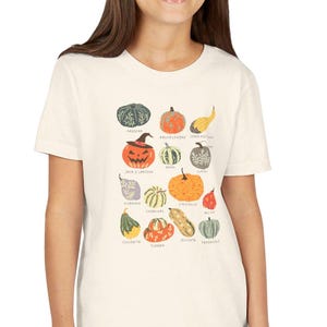Puede incluir: Camiseta color crema con una colorida ilustración de varias calabazas, incluyendo Kabocha, Knucklehead y Jack O'Lantern. Los nombres de cada variedad de calabaza están impresos debajo. Un diseño divertido para el otoño.