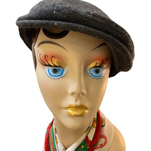 May include: A dark gray tweed newsboy cap with flecks of color. The hat is displayed on a mannequin head with painted features, including blue eyes and gold lips. A patterned scarf is visible.