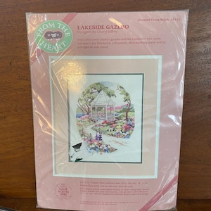 May include: A cross-stitch kit titled "Lakeside Gazebo" designed by Cheryl Jeffrey. The kit features a detailed, colorful scene of a gazebo in a garden setting. The kit includes cotton embroidery thread and 14 count white Aida.