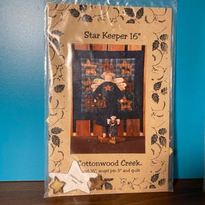 May include: A brown paper package with a printed image of a blue and white quilt with a star-shaped angel holding a basket. The text "Star Keeper 16" and "Cottonwood Creek" is printed on the package.
