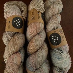 May include: Three skeins of hand-dyed yarn in shades of beige, pink, and blue. The yarn is wrapped around brown paper labels with the text "Hand-dyed with love in Chicago" and the logo "@kraeo kraeo.com".