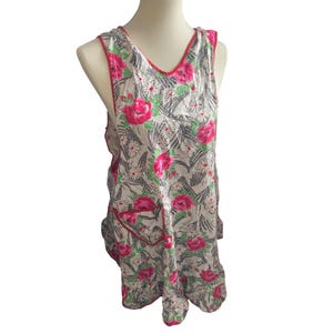 May include: A sleeveless apron with a floral pattern. The apron features large pink roses, white daisies, and green leaves on a light gray background. It has a red trim and a pocket.