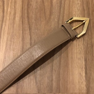 May include: A light brown leather belt with a gold-toned buckle. The buckle is shaped like a pointed oval.