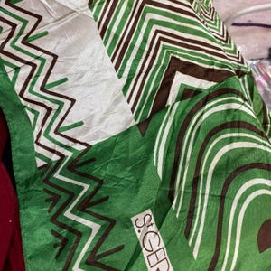 May include: A green, white, and brown patterned silk scarf. The scarf features a repeating geometric design with zigzags and curved lines. The text "S. GER" is visible in the corner. The scarf is made of a shiny, lightweight material.