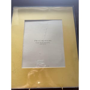 May include: A gold picture frame with a white mat. The mat has the text "Pottery Barn" and "colored mat for wood gallery passe-partouts" printed on it. The frame is 8 x 12 inches (20 x 30 cm).