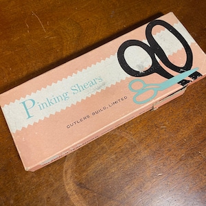 May include: A pink box of pinking shears with the text "Pinking Shears" in light blue. The box has a black and light blue scissors graphic. The text "CUTLERS GUILD, LIMITED" is also visible.