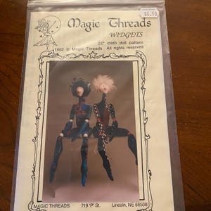 May include: A Magic Threads cloth doll pattern package featuring two seated dolls. The dolls are dressed in black and blue outfits. The package text includes "WIDGETS" and "22" cloth doll pattern".
