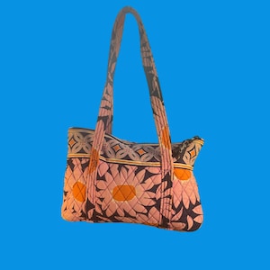 May include: Quilted tote bag with a floral pattern in pink, orange, and navy blue. The bag features a quilted design with large daisy-like flowers. The handles are also quilted and match the bag's pattern. The bag is set against a blue background.