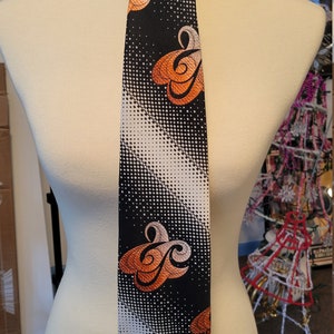 May include: A black tie with a white and orange geometric pattern. The pattern features a repeating design of swirls and dots.