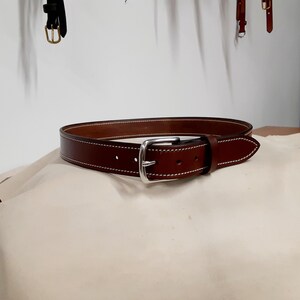 May include: A brown leather belt with a silver buckle. The belt has white stitching along the edges. The belt is resting on a beige surface.