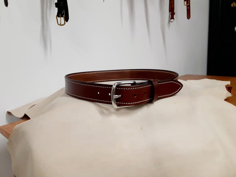 May include: A brown leather belt with a silver buckle. The belt has white stitching along the edges. The belt is resting on a beige surface.