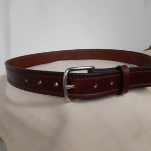 May include: A brown leather belt with a silver-colored buckle. The belt has a classic design with visible stitching and several holes for adjustment. The leather appears to be of high quality.