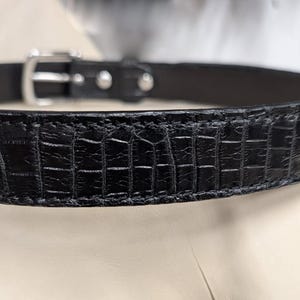 Op de afbeelding: Een zwarte riem met een gestructureerd krokodil-reli&euml;fpatroon over de hele band. De riem is voorzien van een gepolijste zilverkleurige metalen gesp en bijpassende zilverkleurige klinknagels die de band bij de sluiting vastzetten.