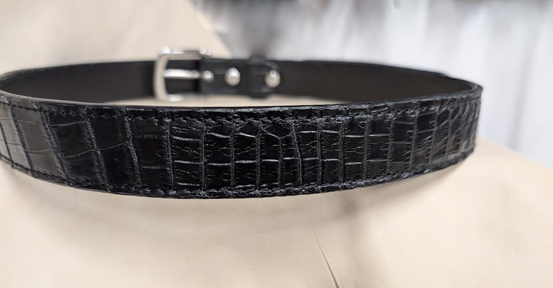 Op de afbeelding: Een zwarte riem met een gestructureerd krokodil-reli&euml;fpatroon over de hele band. De riem is voorzien van een gepolijste zilverkleurige metalen gesp en bijpassende zilverkleurige klinknagels die de band bij de sluiting vastzetten.