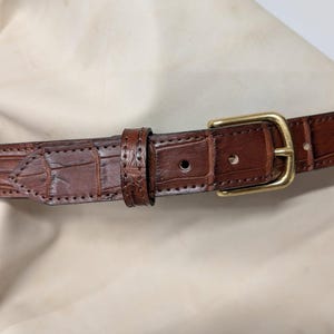 Op de afbeelding: Een bruine leren riem met een textuur van krokodillenleer, voorzien van een gepolijste goudkleurige metalen gesp, een bijpassende leren lus en zichtbare stiksels langs de randen van de riem en het puntige uiteinde van de riem.