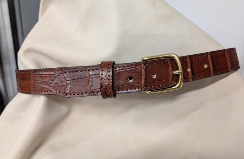 Op de afbeelding: Een bruine leren riem met een textuur van krokodillenleer, voorzien van een gepolijste goudkleurige metalen gesp, een bijpassende leren lus en zichtbare stiksels langs de randen van de riem en het puntige uiteinde van de riem.