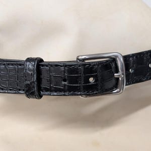 Op de afbeelding: Een zwarte riem met een gestructureerd krokodillenpatroon over de gehele band. De riem is voorzien van een gepolijste zilverkleurige metalen gesp, een bijpassende lederen lus en diverse voorgeponste gaten voor een verstelbare pasvorm.