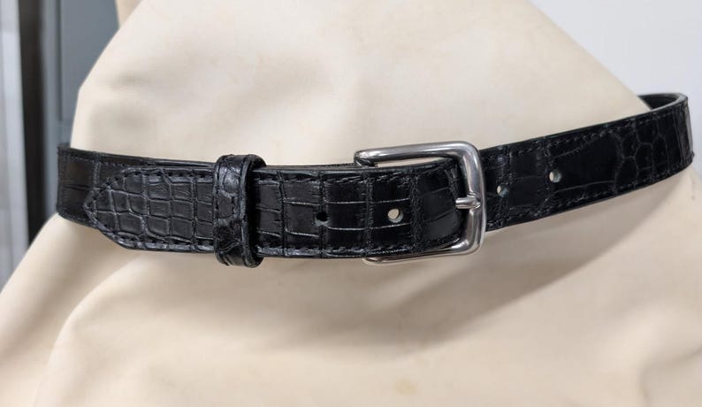 Op de afbeelding: Een zwarte riem met een gestructureerd krokodillenpatroon over de gehele band. De riem is voorzien van een gepolijste zilverkleurige metalen gesp, een bijpassende lederen lus en diverse voorgeponste gaten voor een verstelbare pasvorm.